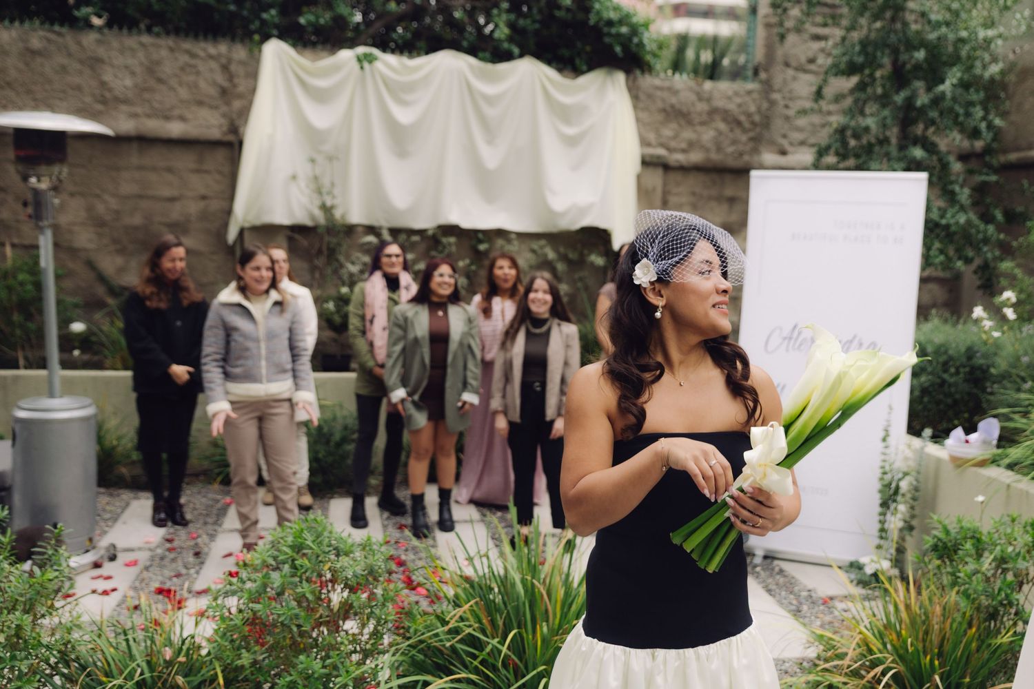 Bride in black strapless dress and birdcage veil holds calla lilies while guests watch at outdoor garden wedding.