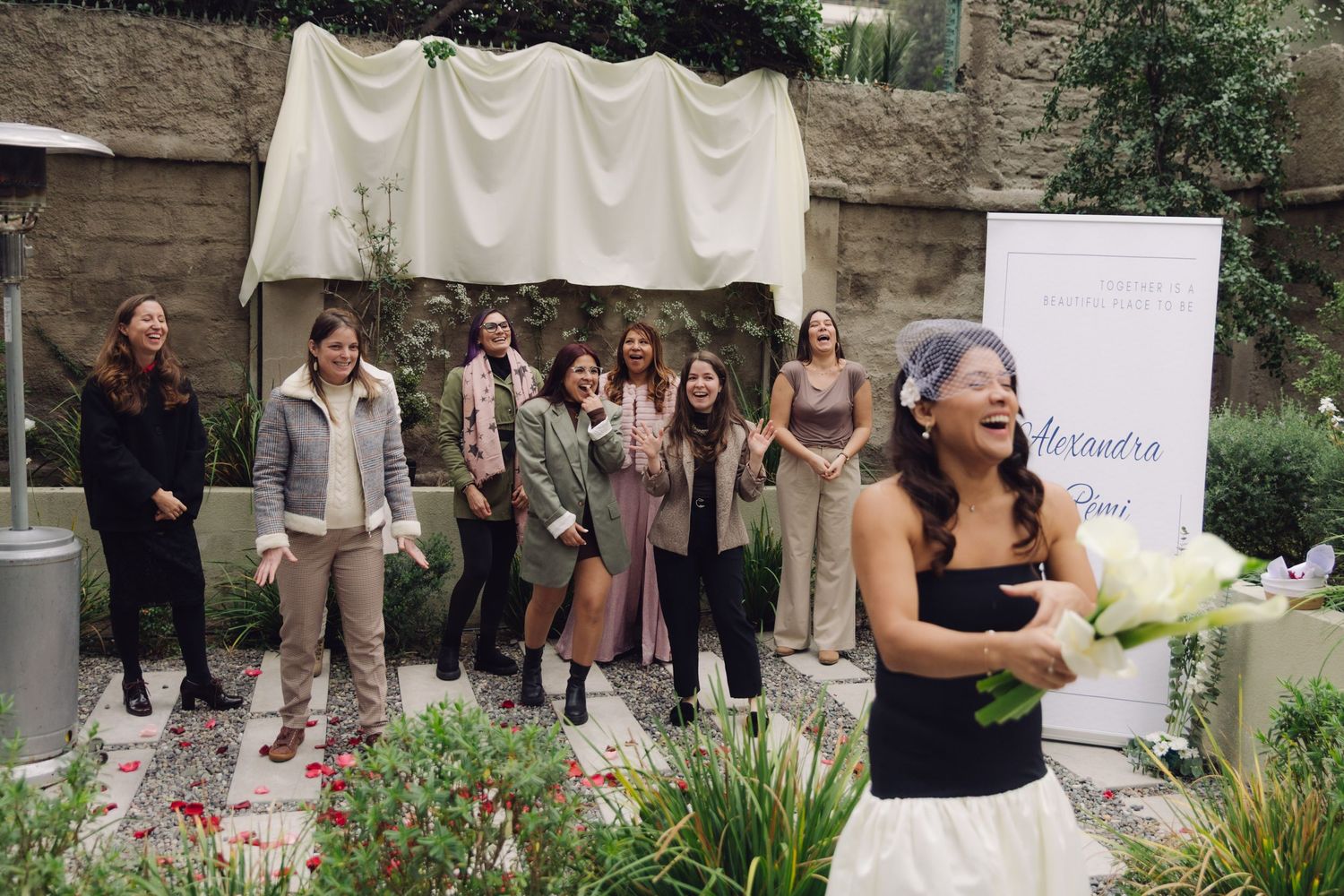 Bride laughing joyfully while tossing bouquet of white calla lilies to excited guests at outdoor garden wedding.