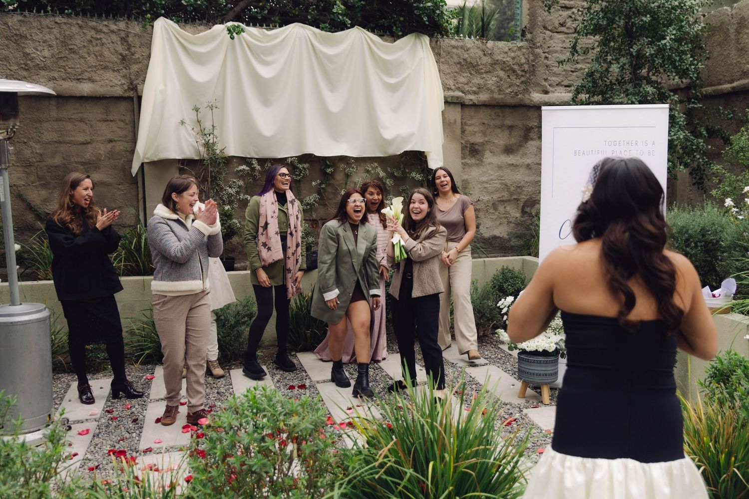 Wedding guests eagerly await bouquet toss from bride in black dress at elegant outdoor garden ceremony.