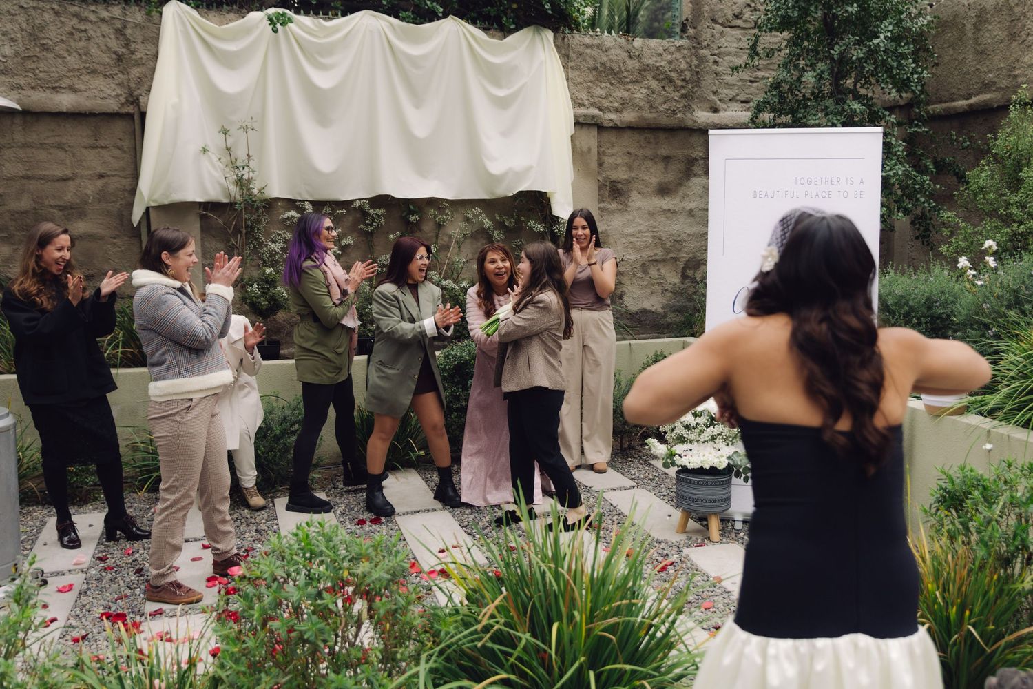 Bride in black strapless gown prepares to toss white bouquet to cheering guests at outdoor garden wedding.