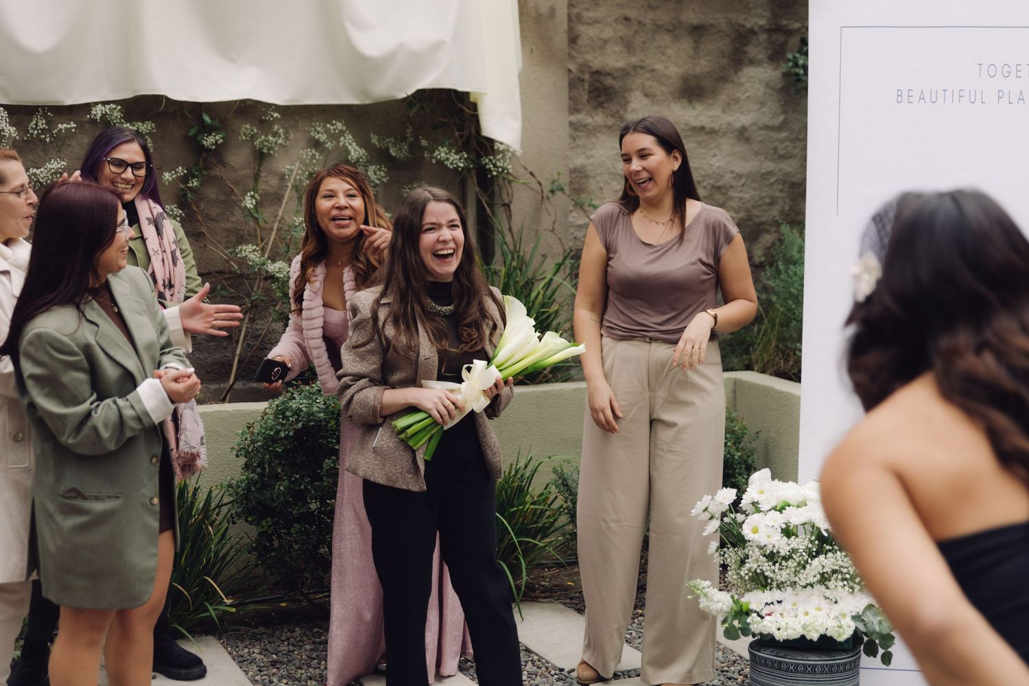 Excited wedding guest catches bridal bouquet of white calla lilies while guests celebrate at outdoor reception.