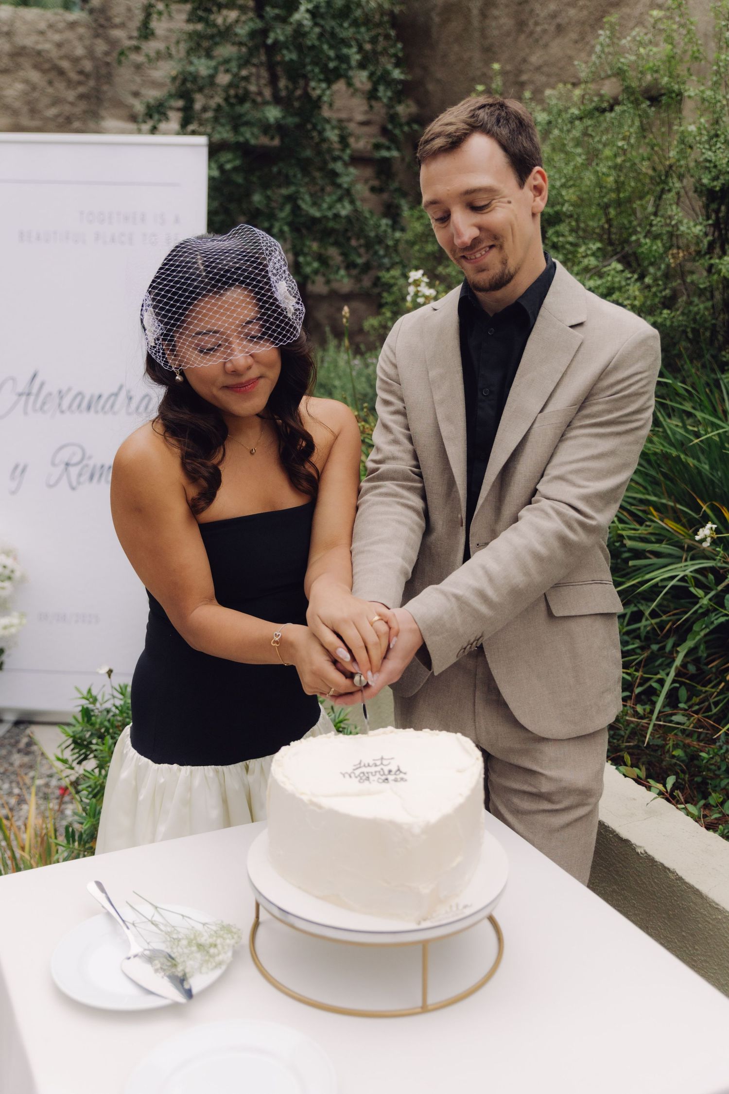 Bride and groom cut heart-shaped white wedding cake together at outdoor garden reception, sign reading Alexandra.