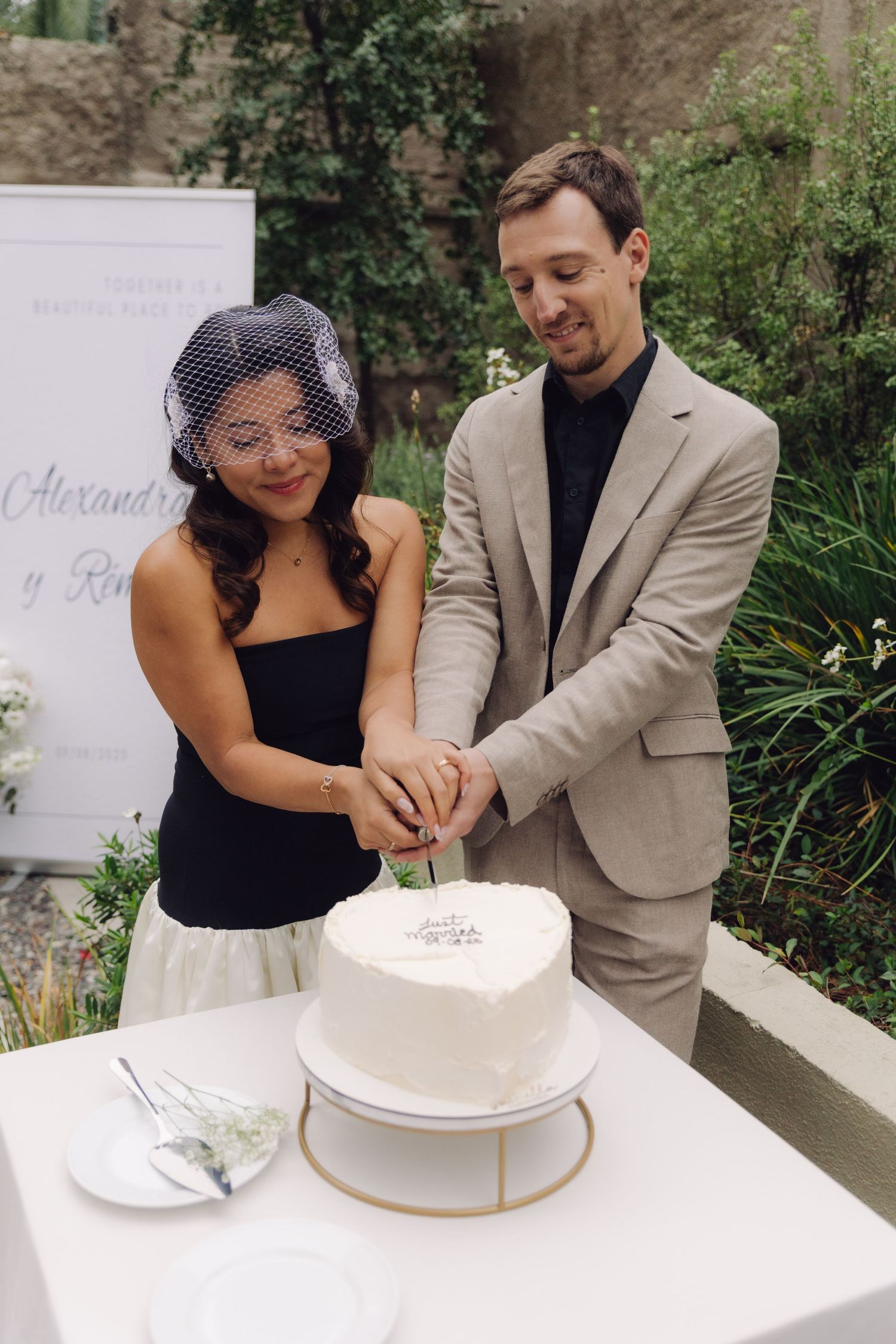 Smiling newlyweds cut elegant heart-shaped white wedding cake at outdoor garden reception, bride wearing birdcage veil.