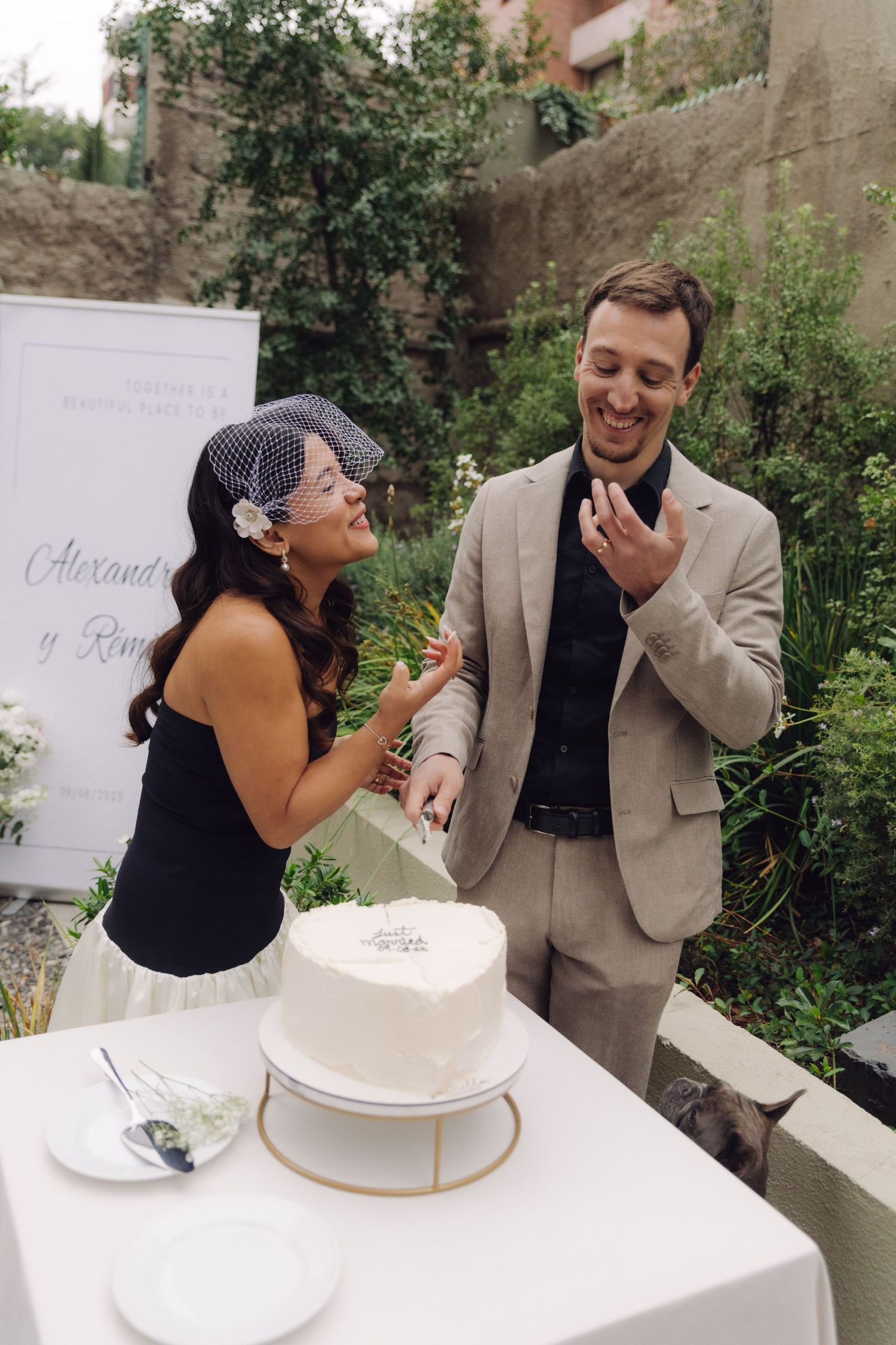 Bride and groom laughing while cutting heart-shaped Just Married wedding cake outdoors.