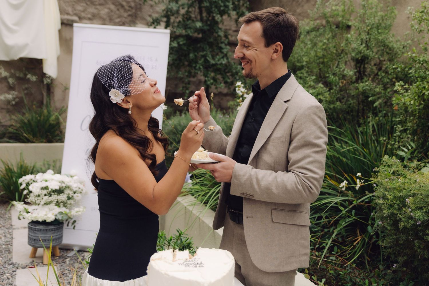 Bride in black dress and birdcage veil feeding groom wedding cake outdoors.