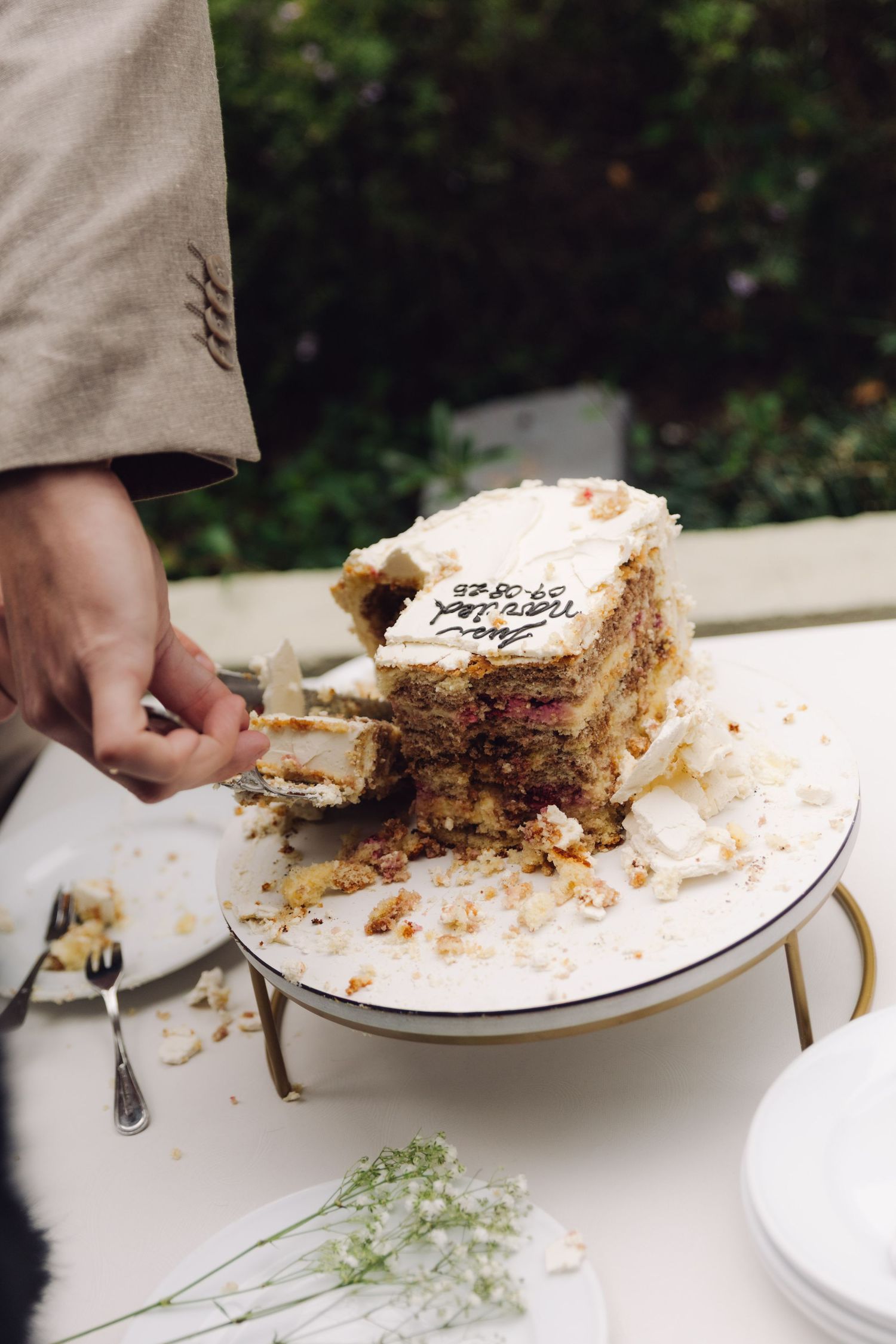 Groom serving slices from a partially cut layered wedding cake on a gold stand.
