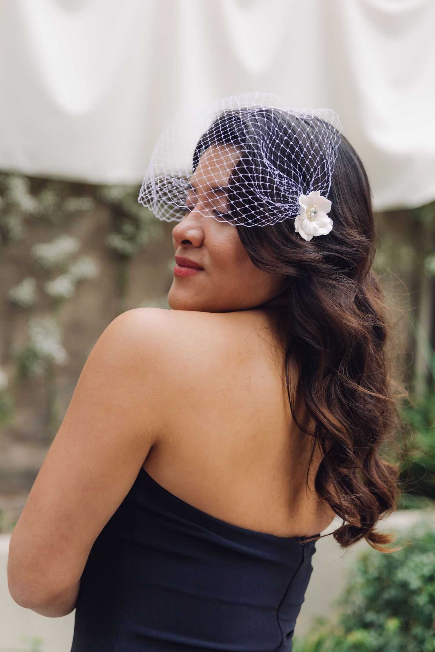 Bride in strapless black dress with birdcage veil looking over her shoulder outdoors.