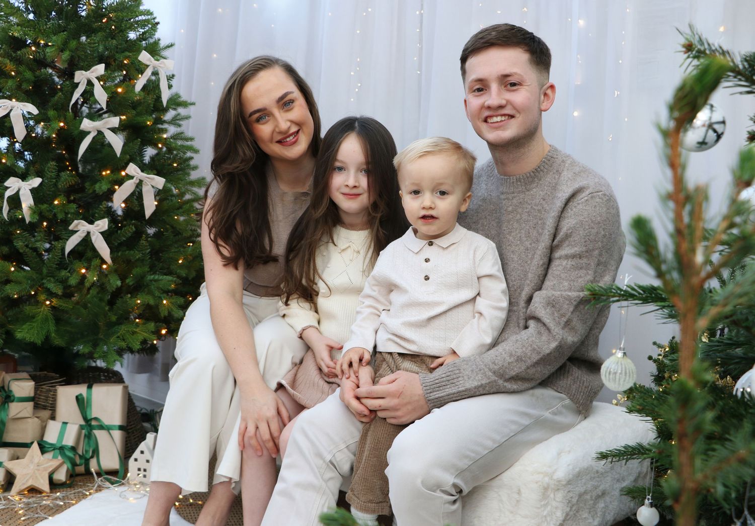 Family poses together in matching cream outfits for holiday photos in front of decorated Christmas tree.