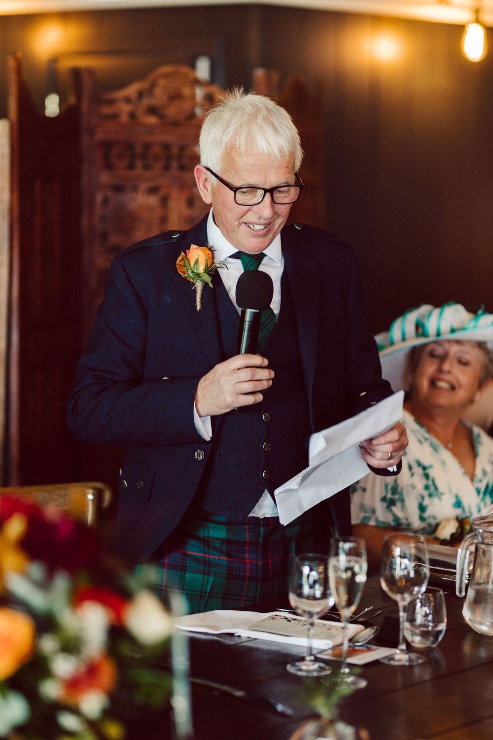 Speaker in tartan kilt and navy blazer gives speech at formal dinner event.