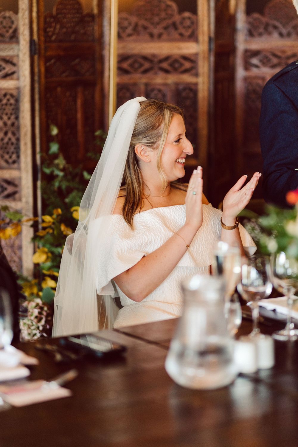 A bride in a white dress and veil laughs expressively during a wedding reception at an ornate venue.