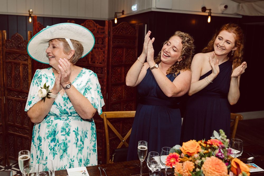 Wedding guests share laughter and conversation during an indoor reception at a rustic venue with warm lighting.