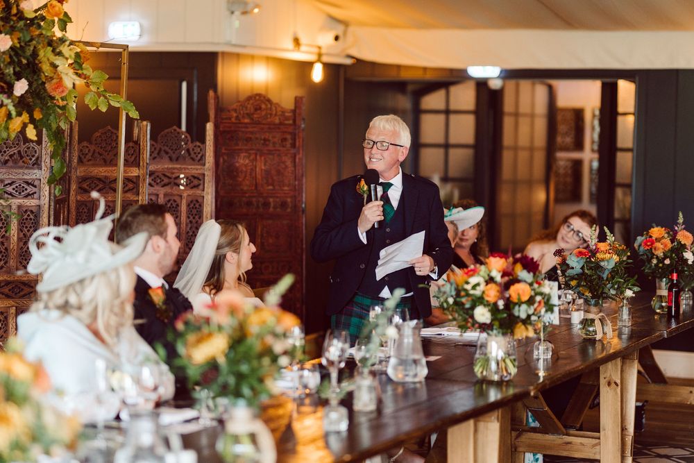 A speaker delivers a toast at an elegant wedding reception with floral centerpieces and warm lighting.