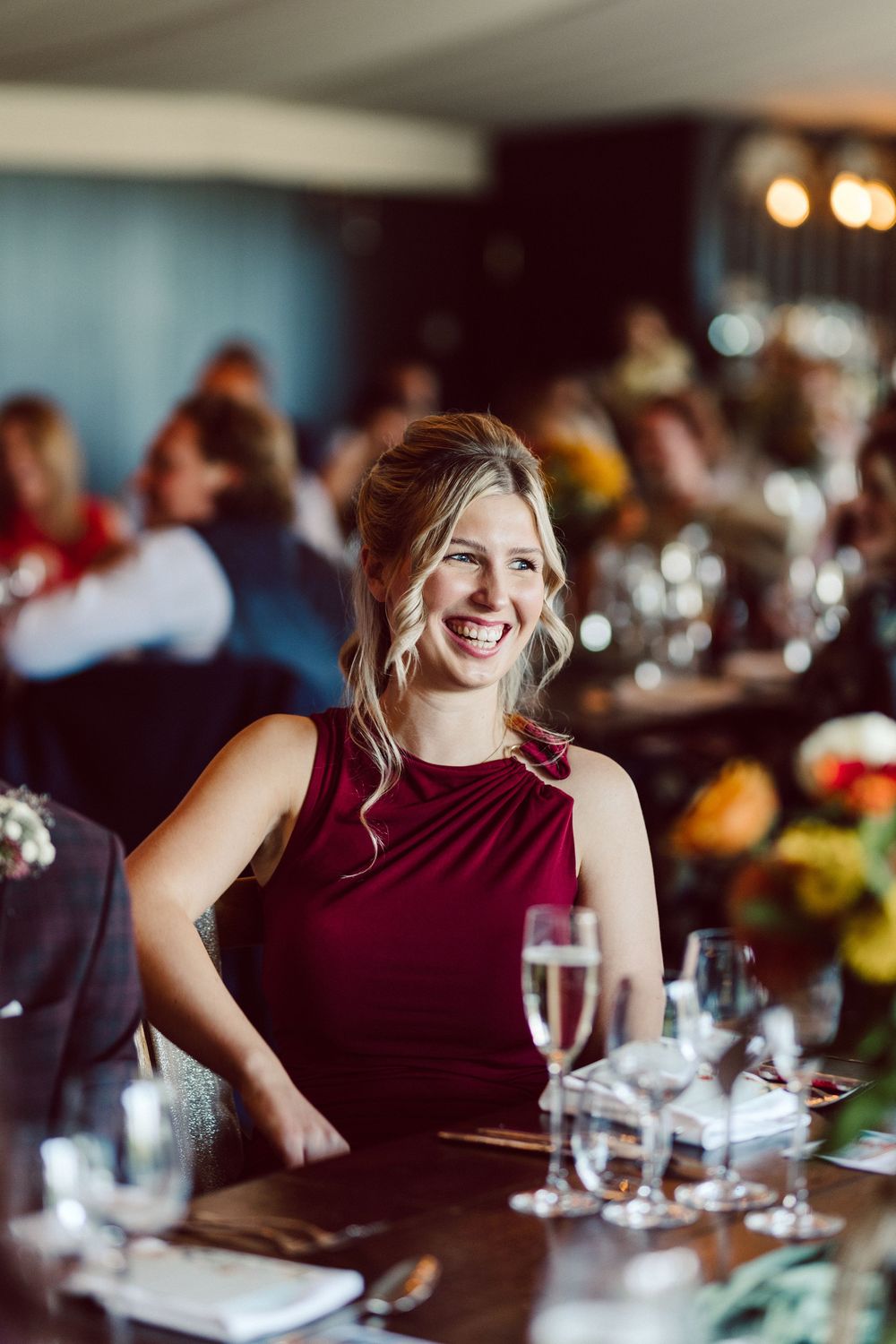 A person in a burgundy dress smiles while dining at a restaurant with soft lighting and table settings.