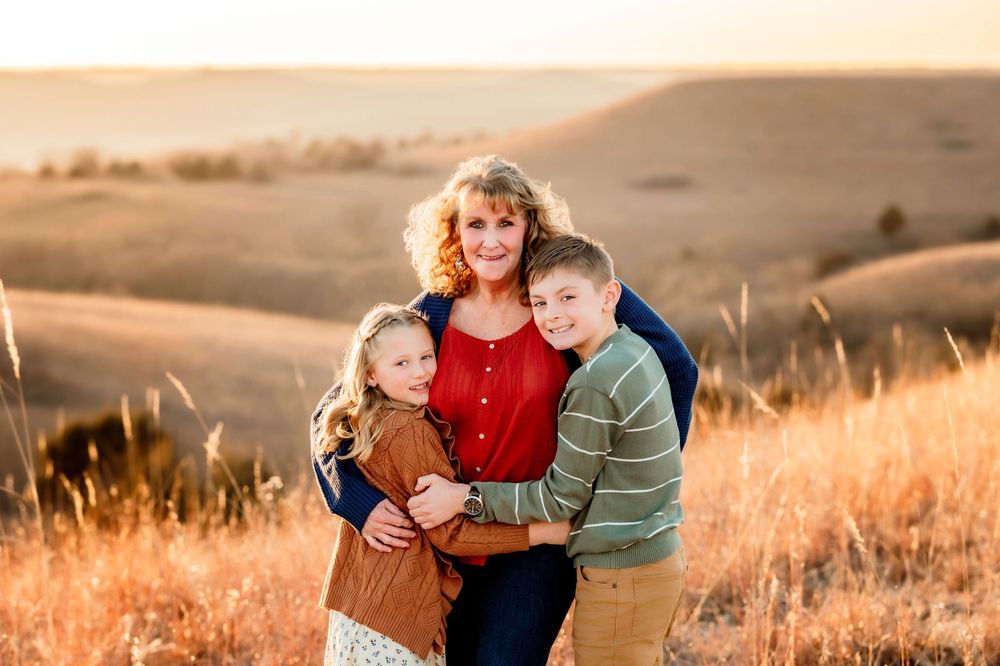 Family embraces together in golden sunlit field during sunset with rolling hills in background.