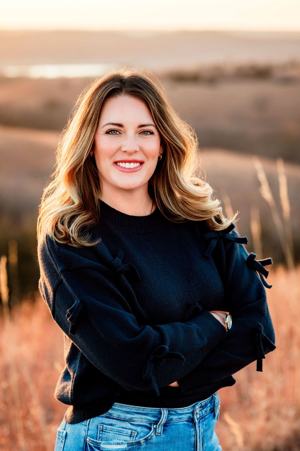 Woman in black ruffled sweater and jeans poses outdoors during golden hour sunset in field.