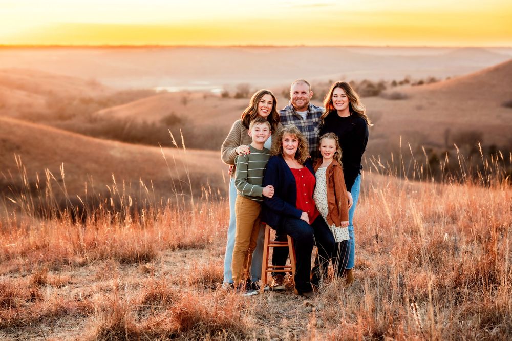 A group of people sit together on a grassy hillside during golden hour, creating a warm and intimate outdoor scene.
