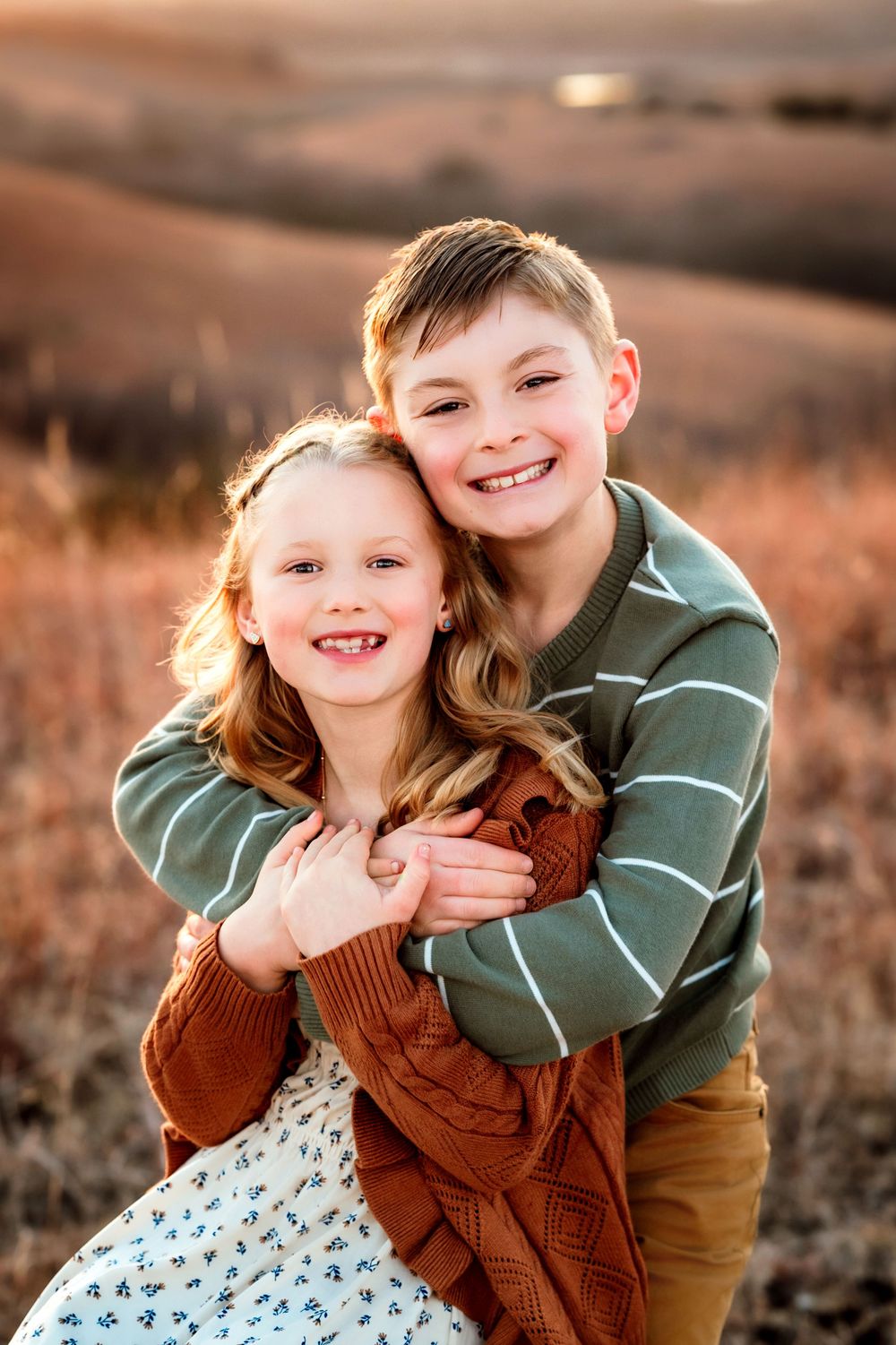 Two siblings share a sweet candid moment in a field at sunset wearing green and brown clothing.