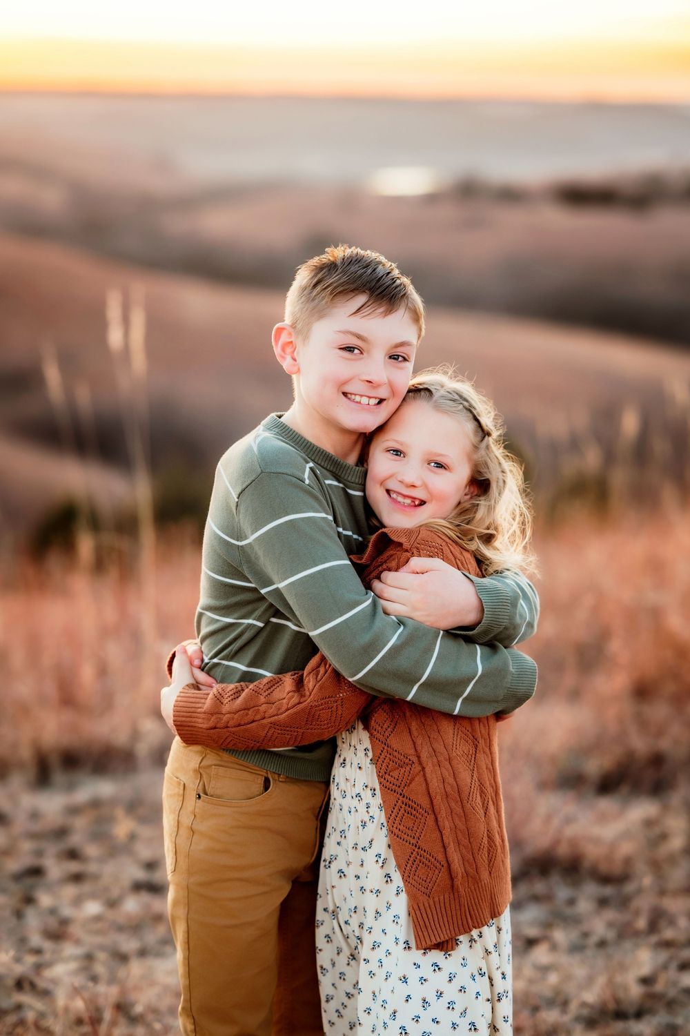 Two children embrace in a warm sunset glow while standing in a golden field during golden hour.