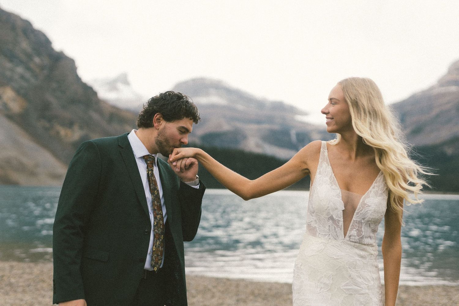 groom kisses bride's hand and she laughs by a mountain lake in Banff
