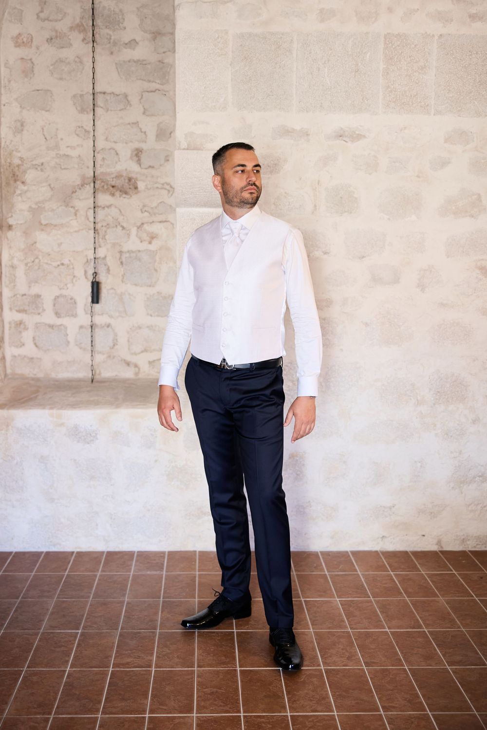 A person in a white shirt and dark pants walking across a room with stone walls and terracotta flooring.