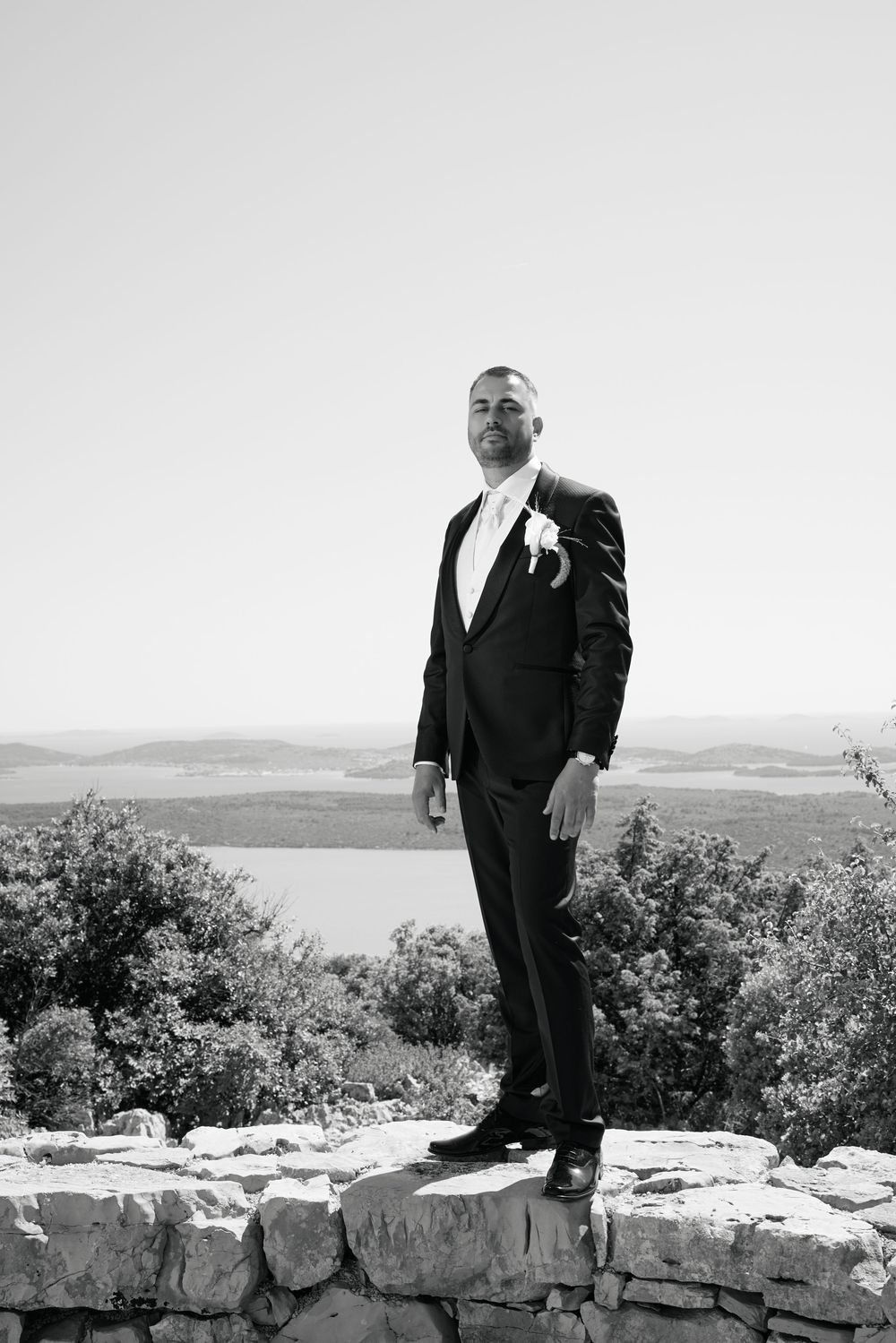 A groom in a black tuxedo stands on a stone wall overlooking a scenic landscape in black and white photography.