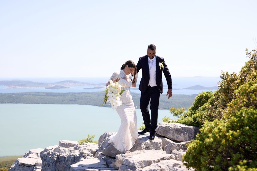 A couple in wedding attire poses on a scenic mountain overlook with water views in the background.