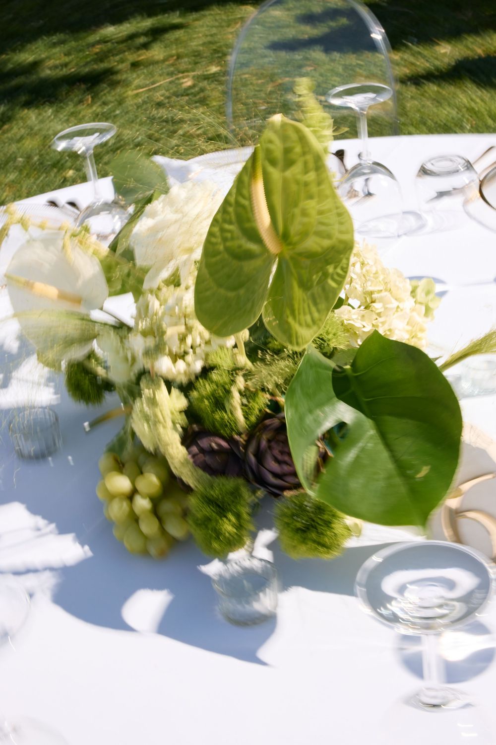 Elegant table setting with white linens features a centerpiece of green anthurium leaves, grapes and hydrangeas in natural light.