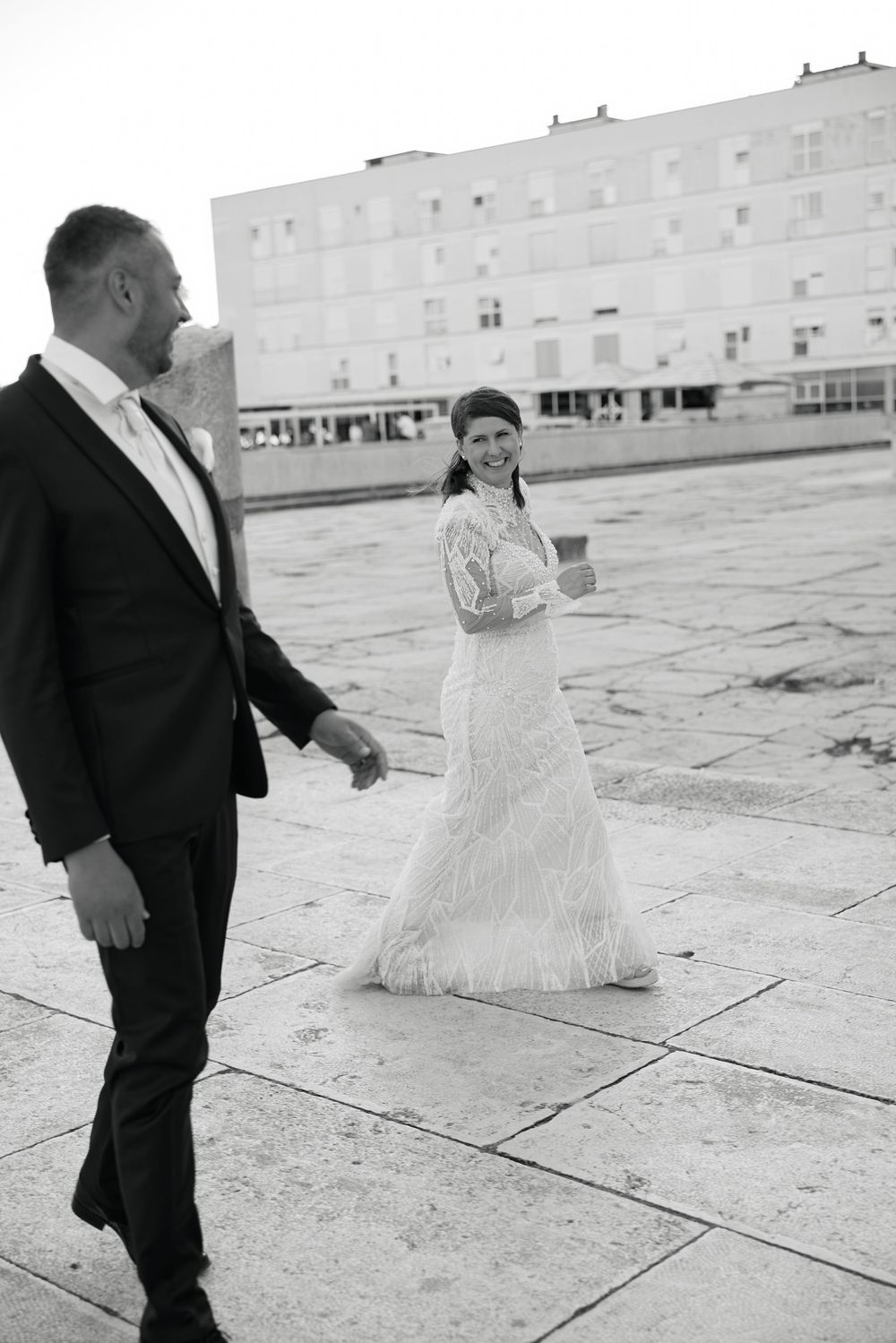A couple in wedding attire poses on an urban plaza with a modern building backdrop.