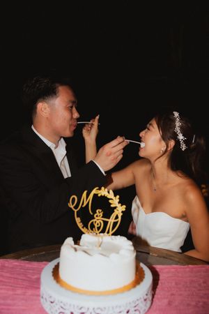 Couple feeding each other cake at their wedding reception with a white decorated cake in the foreground.
