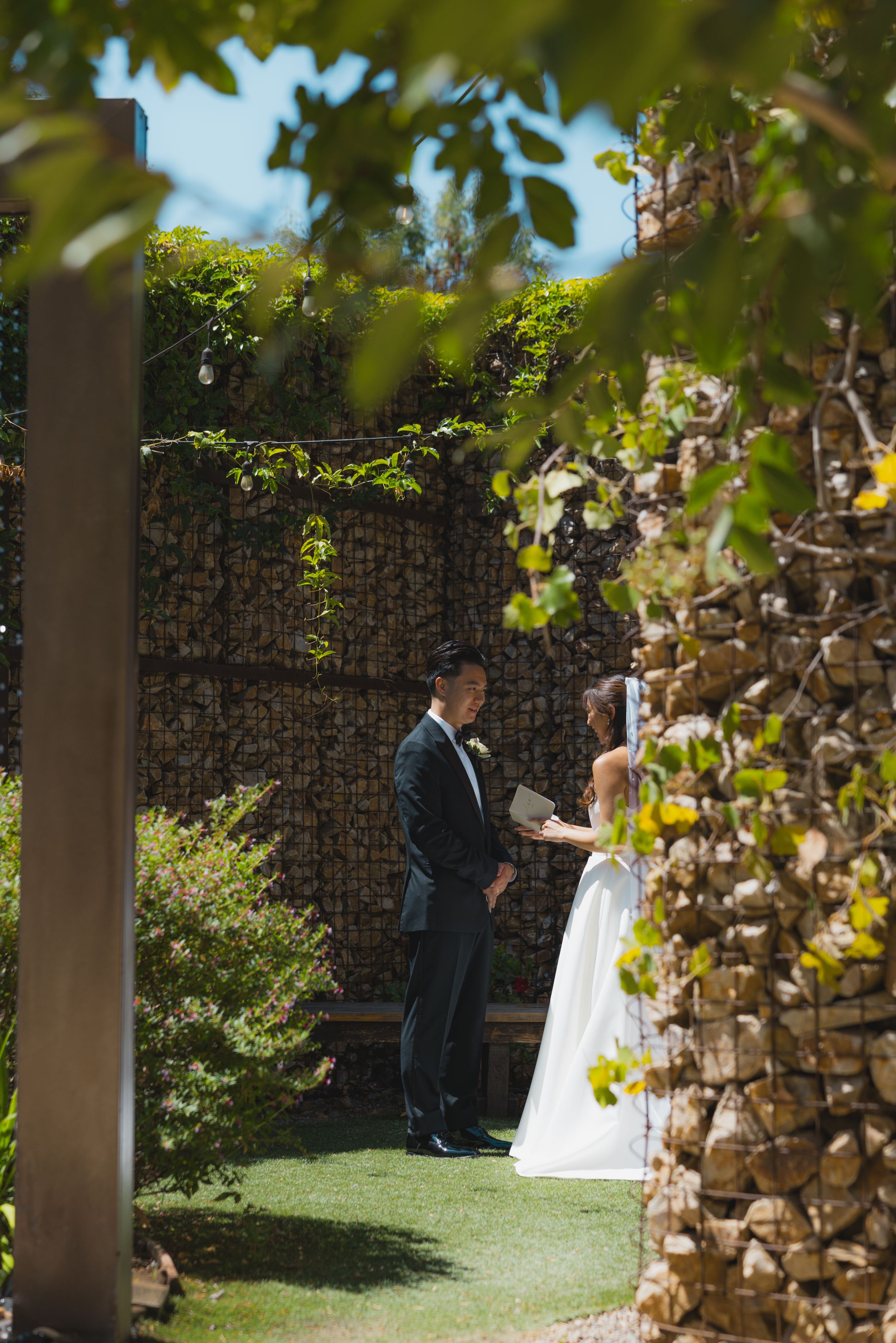 A couple shares an intimate moment in a lush garden setting with stone walls and greenery on a sunny day.