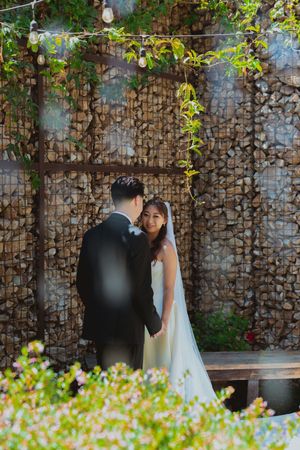 Couple shares intimate moment against stone wall with hanging lights and green vines.