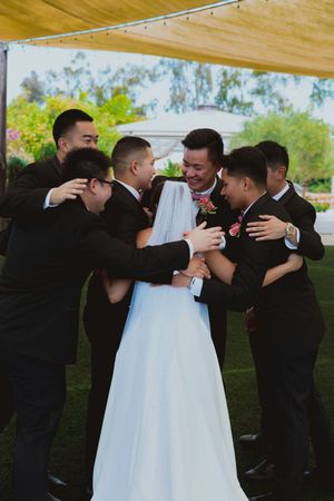Wedding party members share a joyful group hug in formal black suits and white dress at an outdoor ceremony.