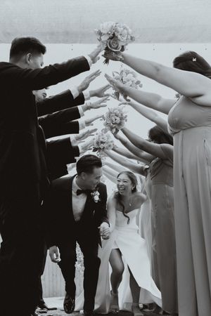 Wedding party forms a tunnel with bouquets as newlyweds duck underneath in black and white photography.
