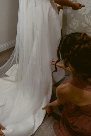 A bridesmaid in a burgundy dress helps adjust the flowing white train of a wedding gown.