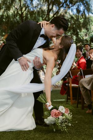 A romantic wedding couple shares a kiss while dipping on a grassy lawn during their outdoor ceremony.