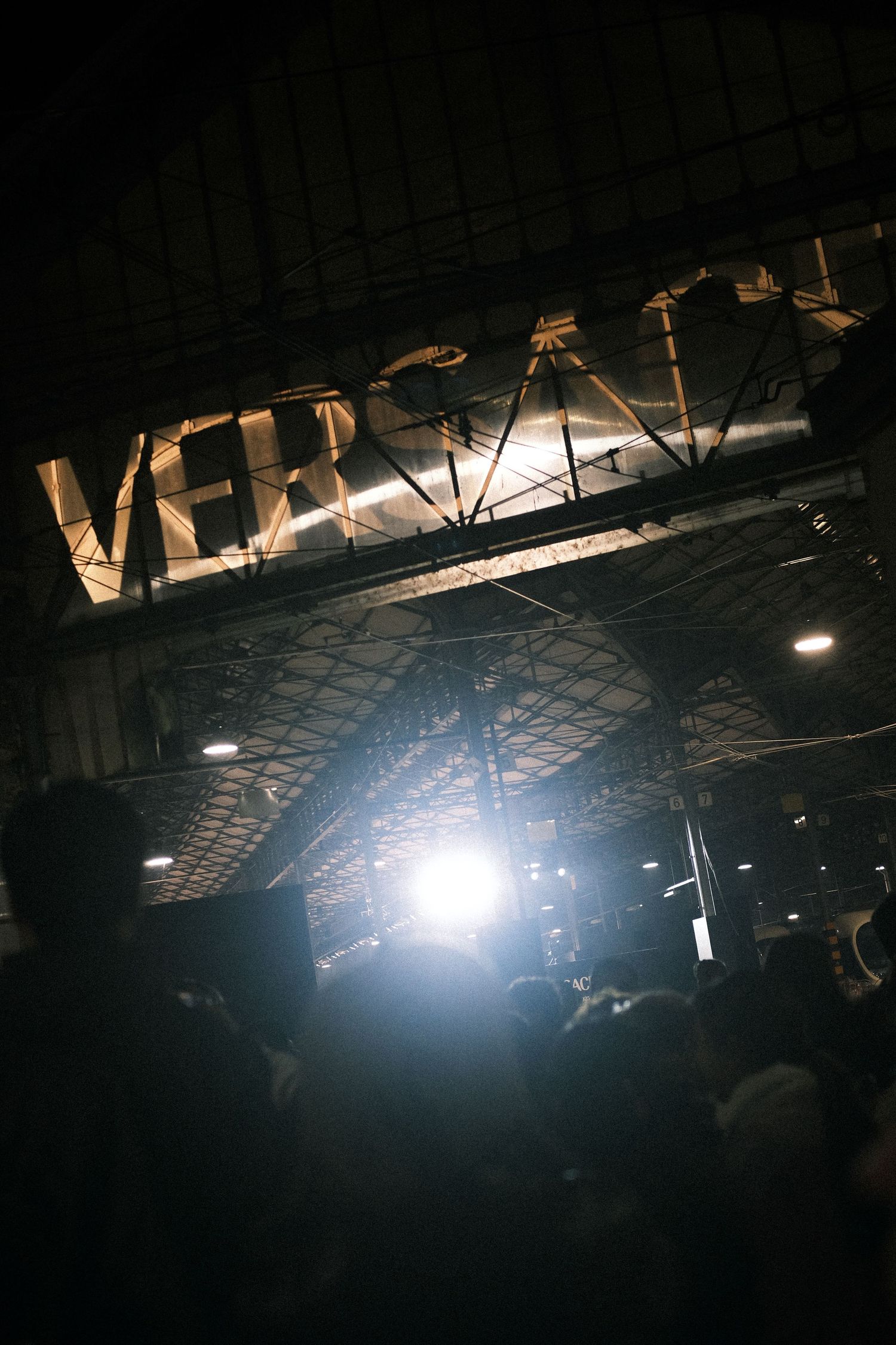Dark concert venue interior with dramatic stage lighting and rigging equipment visible in the ceiling.