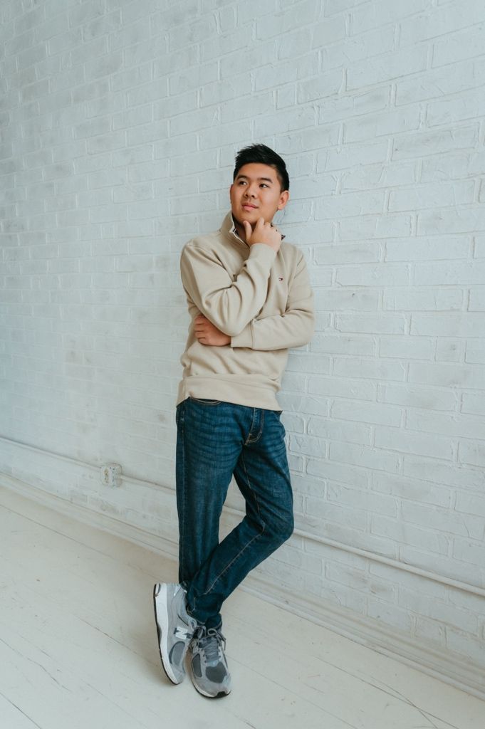 Person in beige sweater and jeans strikes casual pose against white brick background.