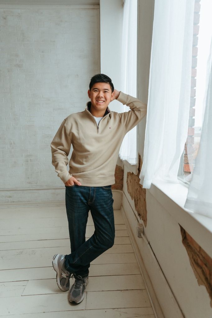 A person in a beige sweater and dark pants poses playfully in a bright white hallway.