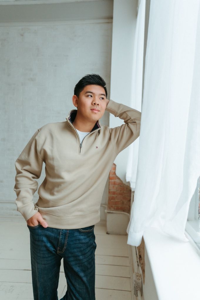 Person in beige sweater and dark jeans poses casually against white wall near window in minimalist setting.