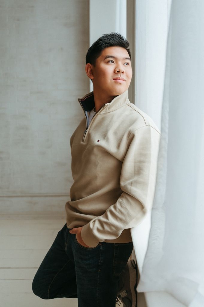 Figure in neutral colored pullover leans playfully against bright window in brick-walled room.