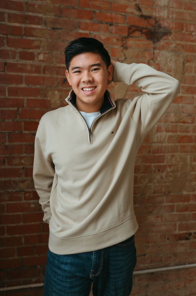 Person in beige quarter-zip sweater and jeans poses casually against brick wall in studio setting.