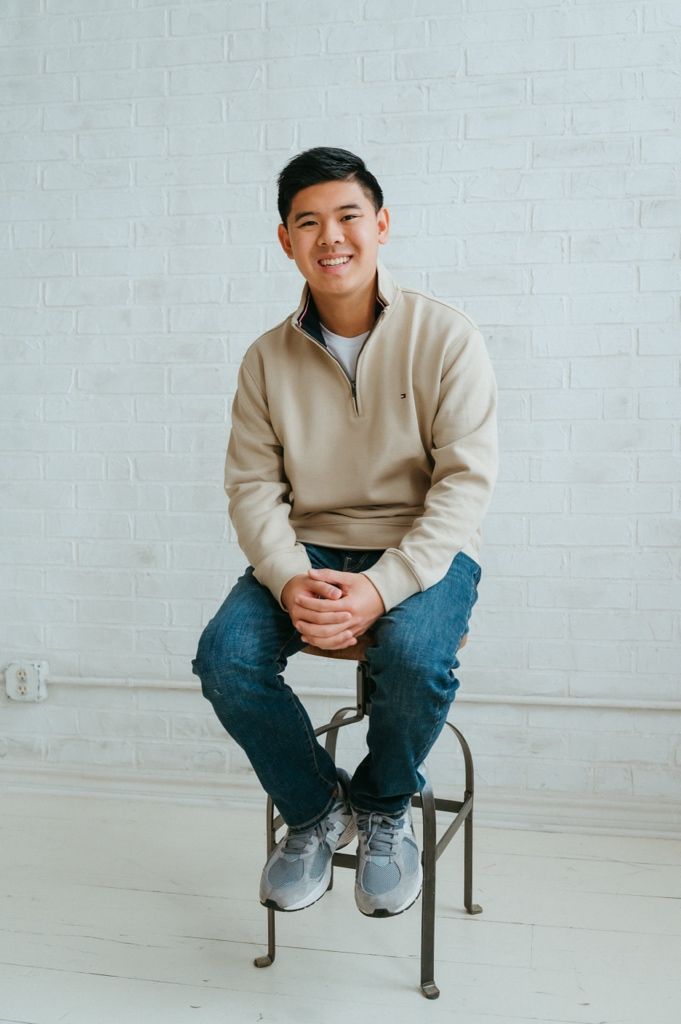 A person in a beige sweater and jeans sits casually on a chair against a white brick wall.