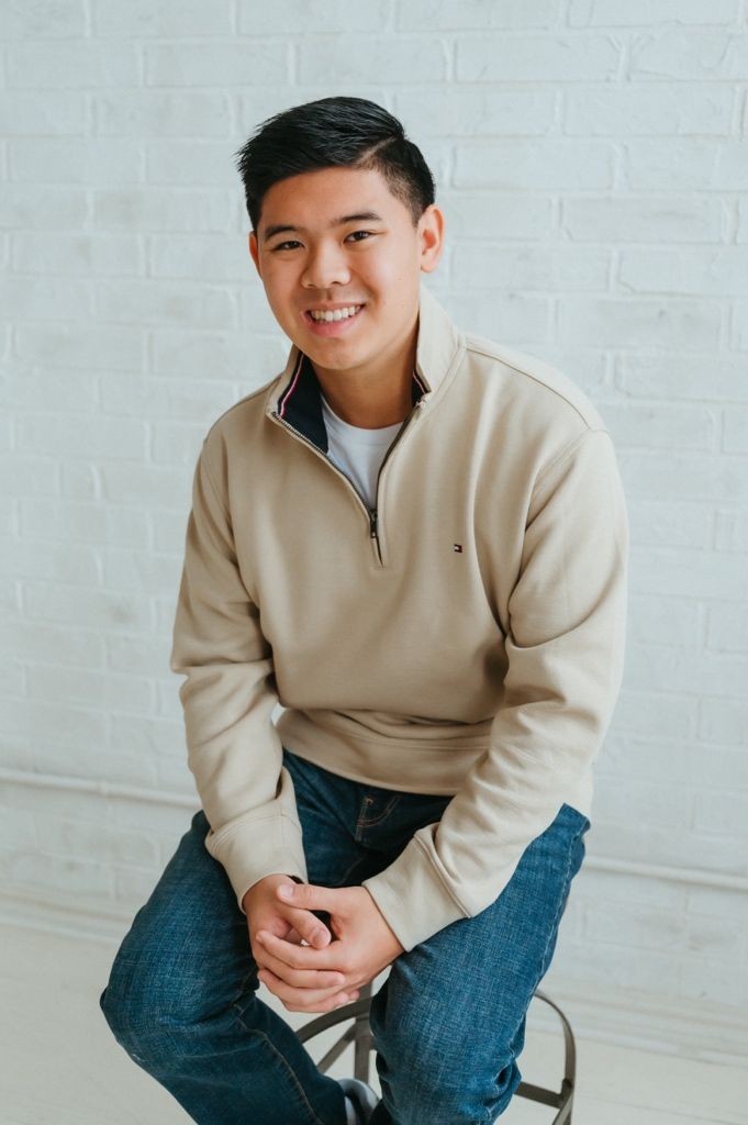 Person in beige quarter-zip sweater and blue jeans sits casually on chair against white brick wall.