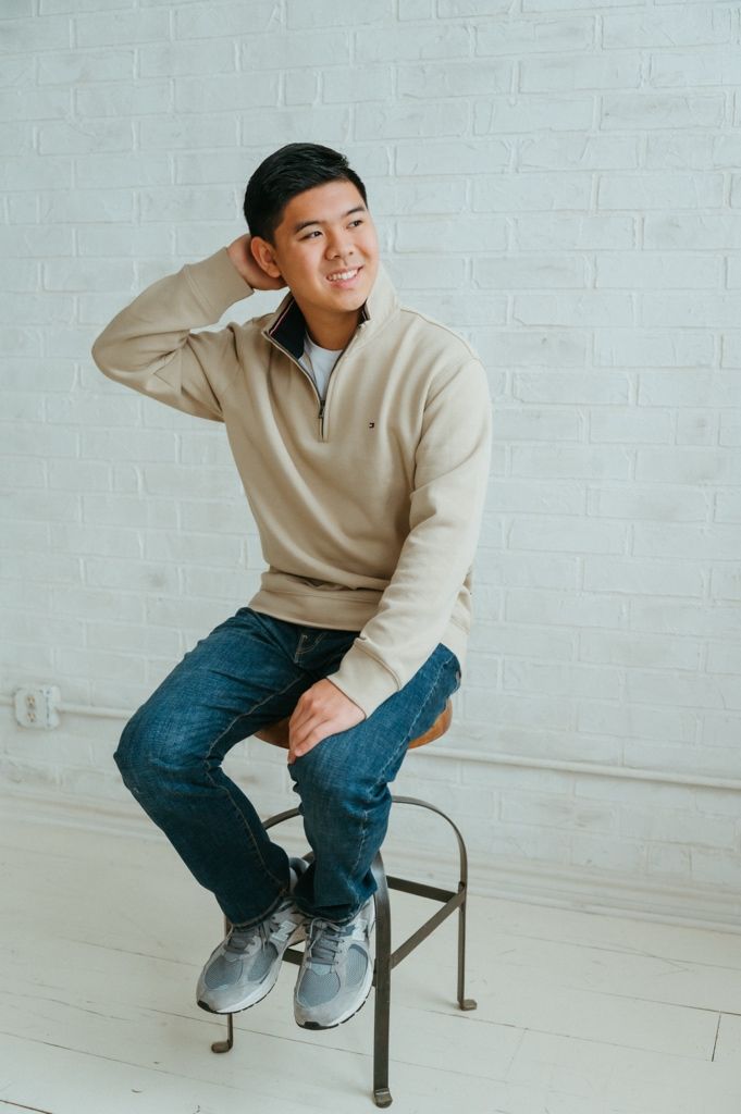 Someone in a beige quarter-zip sweater and jeans sits casually on a metal stool against a white brick wall.