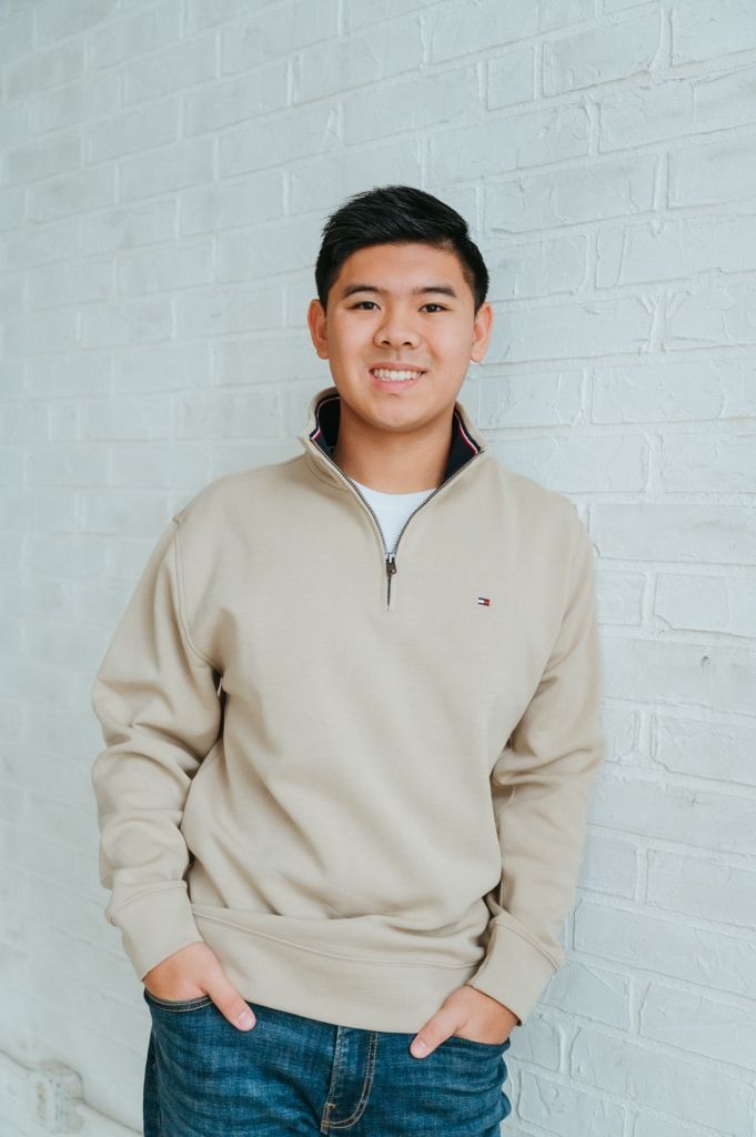 A person in a beige quarter-zip sweater and blue jeans poses against a white brick wall in a casual portrait series.