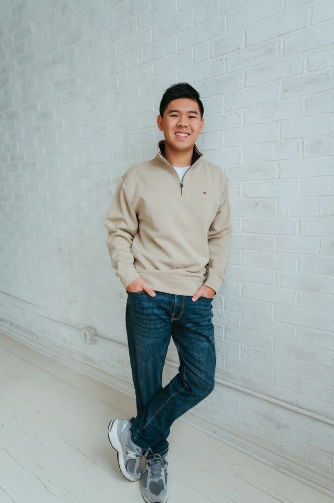 Person in beige sweatshirt and jeans poses casually against white brick wall.