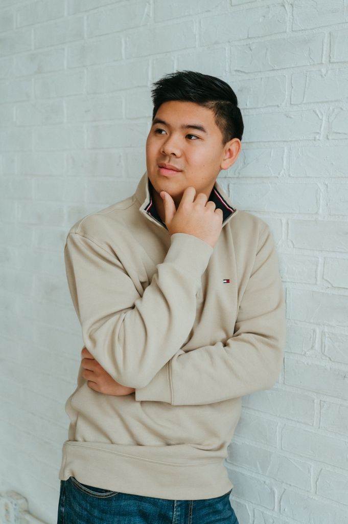 Person wearing beige sweatshirt leans against white brick wall with hand on chin.