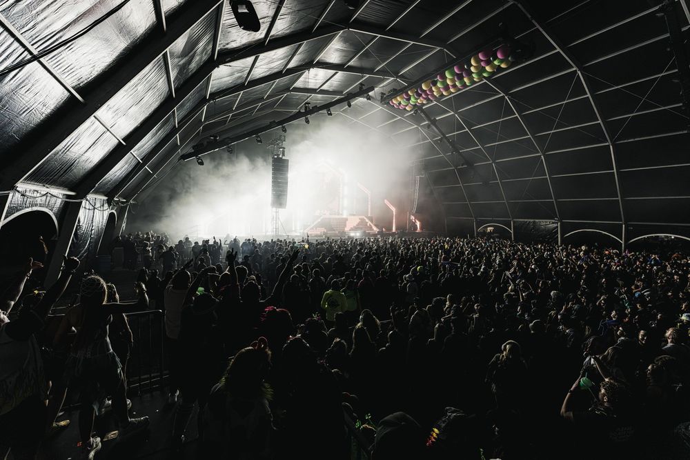 Silhouetted crowd of concert attendees in a dark arena with dramatic lighting and stage effects in the background.