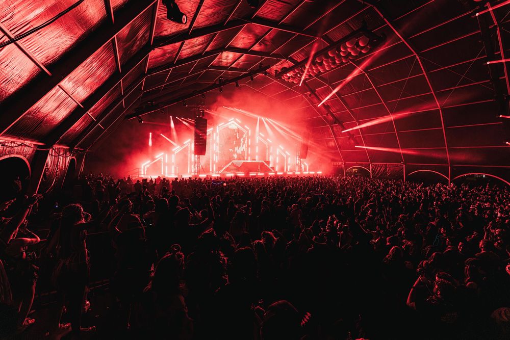 A large concert crowd silhouetted against bright red stage lighting under an arched venue roof during a performance.