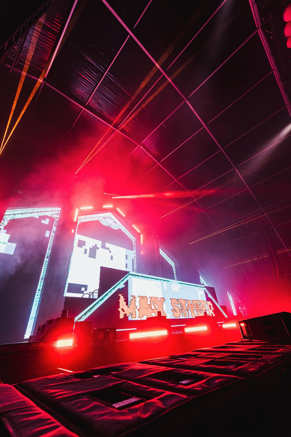 Dramatic red and white stage lighting illuminates a dark concert venue with geometric architectural elements.